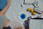 Person lying on the ZAZZA blue picnic blanket, with the picnic collection, styled  with lemons and a plate of blue ice lolly