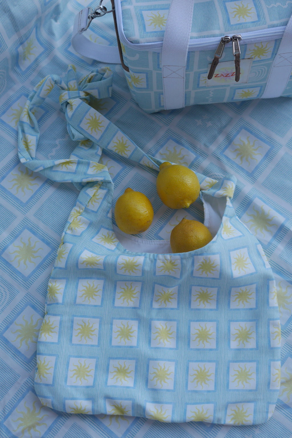 Flat lay image of ZAZZA Blue tote bag with lemons on a matching fabric background (picnic blanket) & other products from the picnic collection