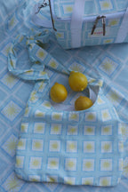 Flat lay image of ZAZZA Blue tote bag with lemons on a matching fabric background (picnic blanket) & other products from the picnic collection