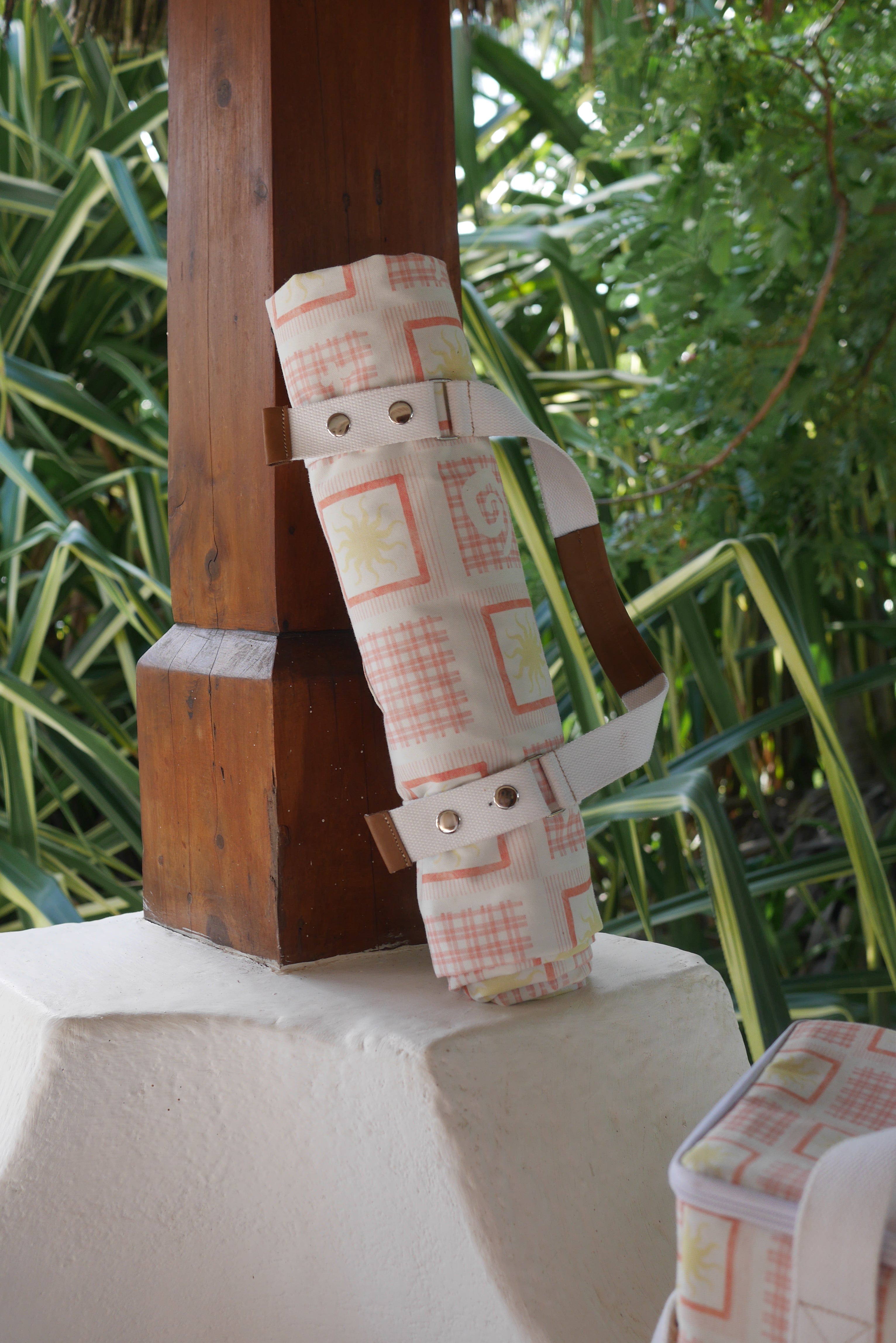 ZAZZA red picnic blanket, rolled &  to a wooden surface, with greenery in the background
